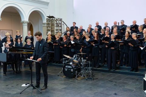 Chorleiter Nico Köhs steht am Podium, der Philharmonische Chor in Gesangsaufstellung hinter ihm auf der Bühne.
